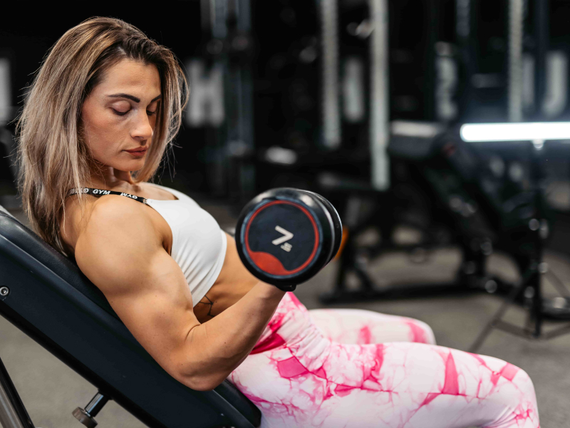 Eine Frau sitzt auf einer Trainingsbank und macht konzentriert Bizeps-Curls mit einer 7-Kilo-Kurzhantel.