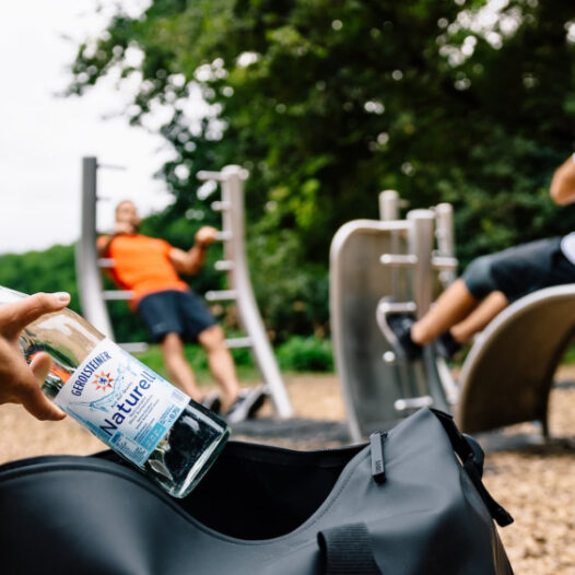 Eine Frau packt eine Flasche Gerolsteiner Wasser mit Mineralstoffen in ihre Tasche, während im Hintergrund zwei Männer ihre Muskeln an Outdoor-Sportgeräten im Park trainieren.