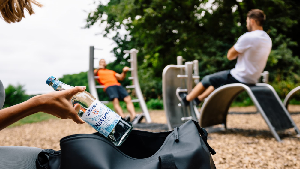 Eine Frau packt eine Flasche Gerolsteiner Wasser mit Mineralstoffen in ihre Tasche, während im Hintergrund zwei Männer ihre Muskeln an Outdoor-Sportgeräten im Park trainieren.