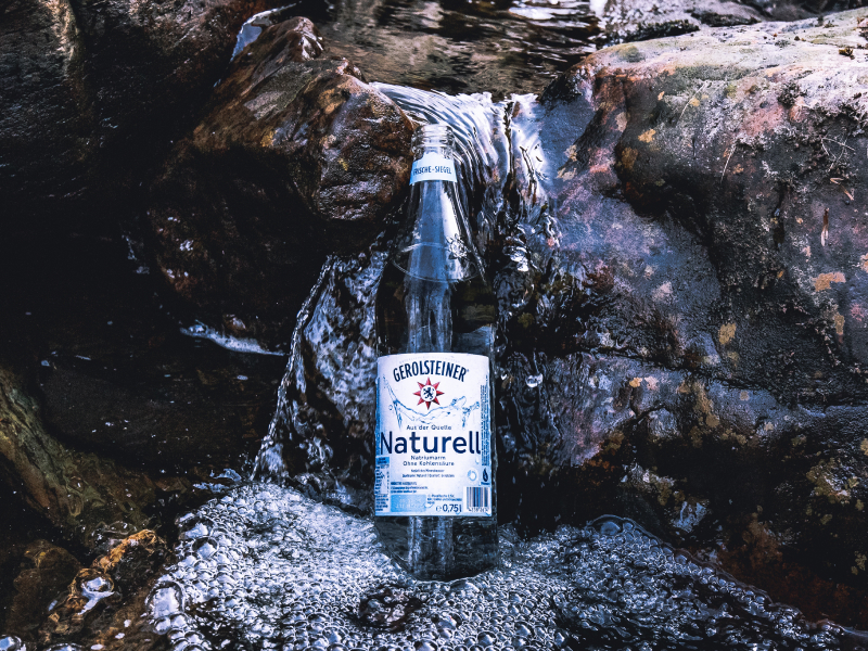 Eine Gerolsteiner Naturell Glasflasche steht auf einem Felsvorsprung eines Bachverlaufs in der Eifel.