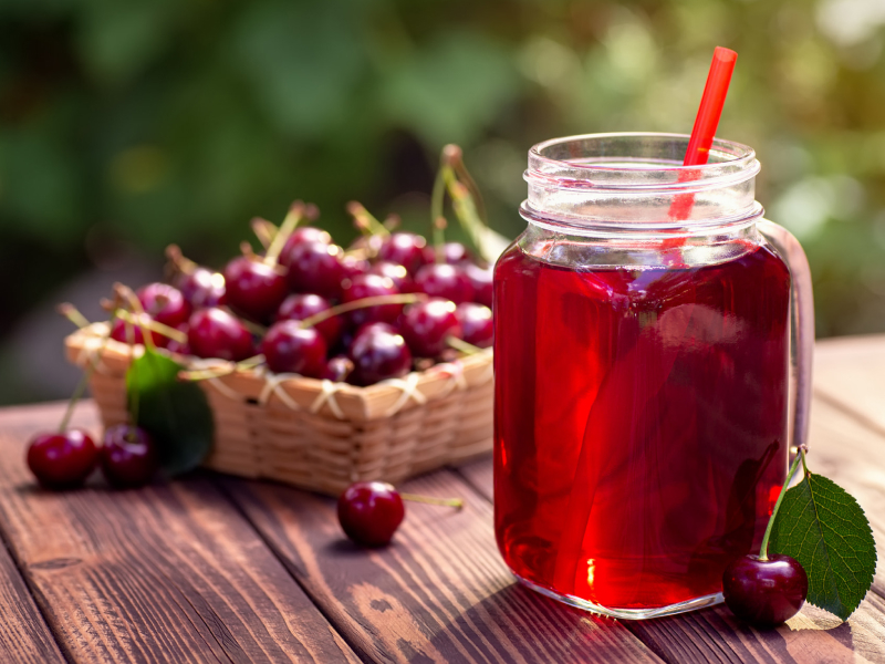 Kirschsaft in einem Einmachglas mit Strohhalm und einer Schale reifer Kirschen im Hintergrund auf einem Holztisch im Freien.