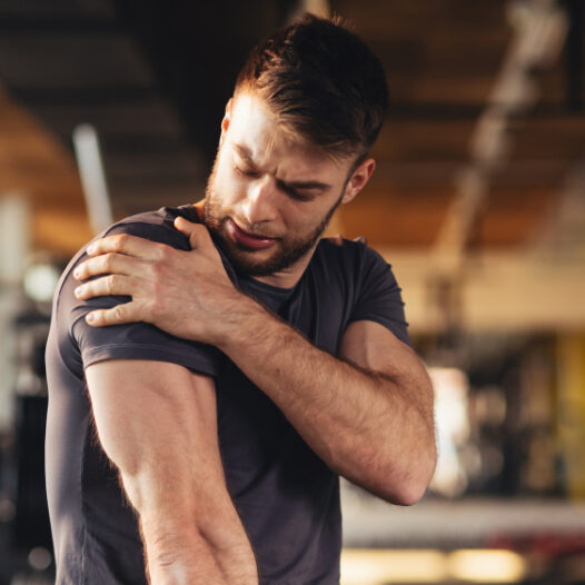 Junger Mann in Sportkleidung sitzt im Fitnessstudio und hält sich die durch Muskelkater schmerzende Schulter.