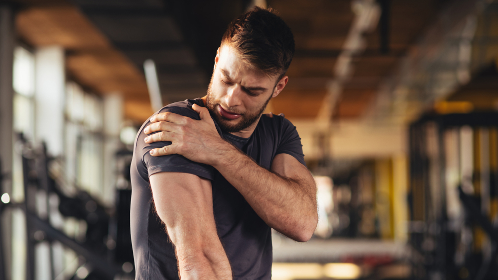 Junger Mann in Sportkleidung sitzt im Fitnessstudio und hält sich die durch Muskelkater schmerzende Schulter.