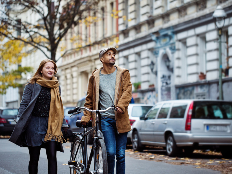 Zwei Freunde, eine Frau und ein Mann in warmer Kleidung, gehen in der Stadt spazieren, um ihren Muskelkater zu lockern. Der Mann schiebt dabei ein Fahrrad.