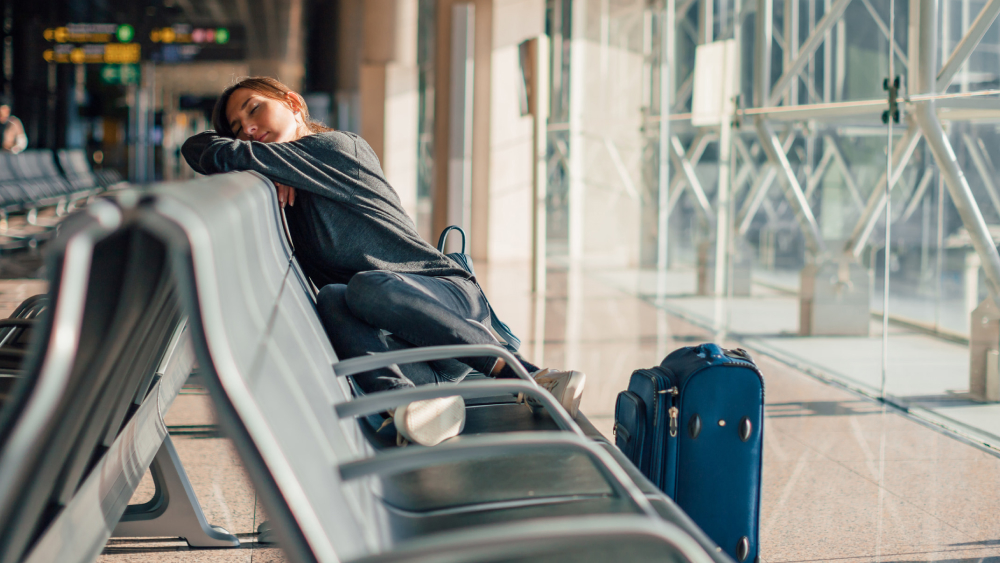 Müde Frau schläft auf Sitzbank am Flughafen. Neben ihr steht ein blauer Koffer.