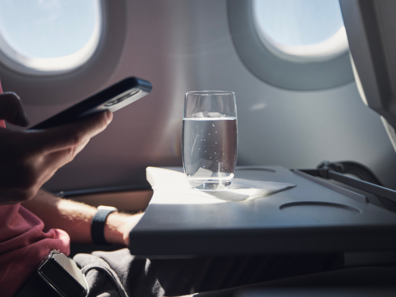 Eine Person sitzt im Flugzeug am Handy. Vor ihr auf dem ausgeklappten Tisch steht ein Glas mit Wasser.